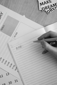 Black and white photo of hand writing 'plan' in notebook with planning materials around.